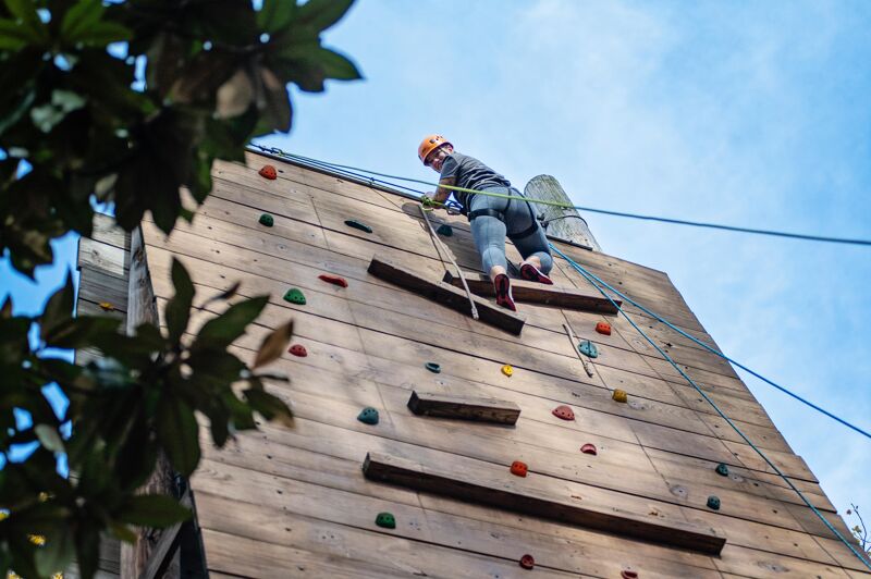 A person wearing a helmet is climbing a wooden climbing wall. The wall has various handholds and footholds attached to it. The person is secured with a rope and harness. The sky is visible in the background. The climbing wall appears to be outdoors, possibly in a park or recreational area.